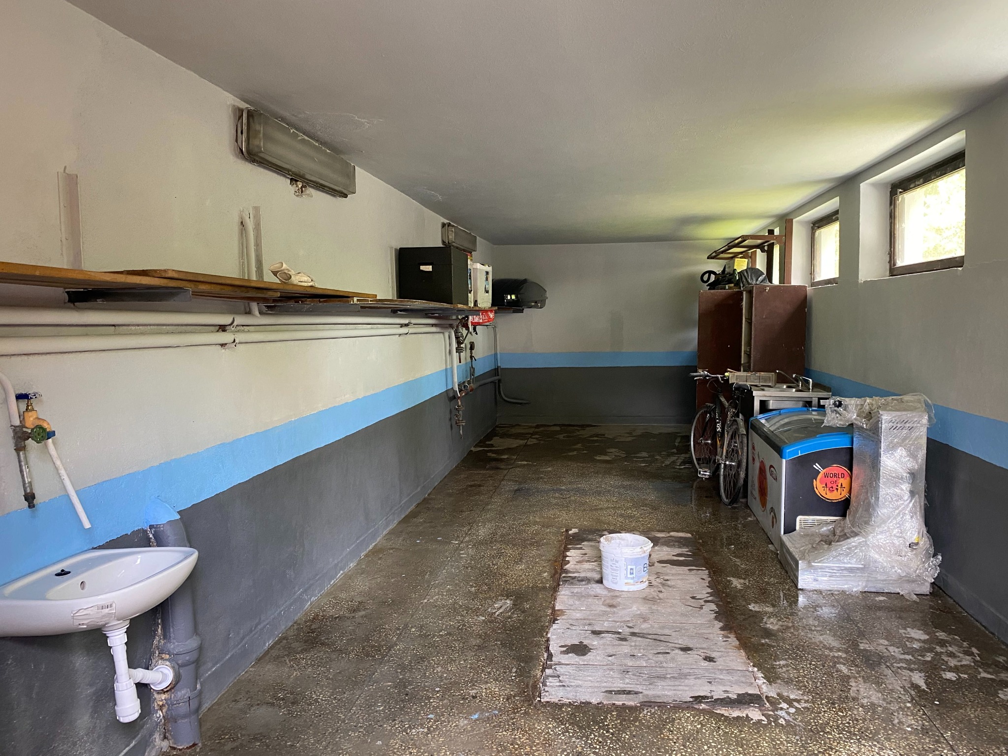 Piwnica po malowaniu ścian na szaro-biało z niebieskim pasem. Widoczna umywalka, okna i sprzęty. Na podłodze stoi wiadro z farbą.