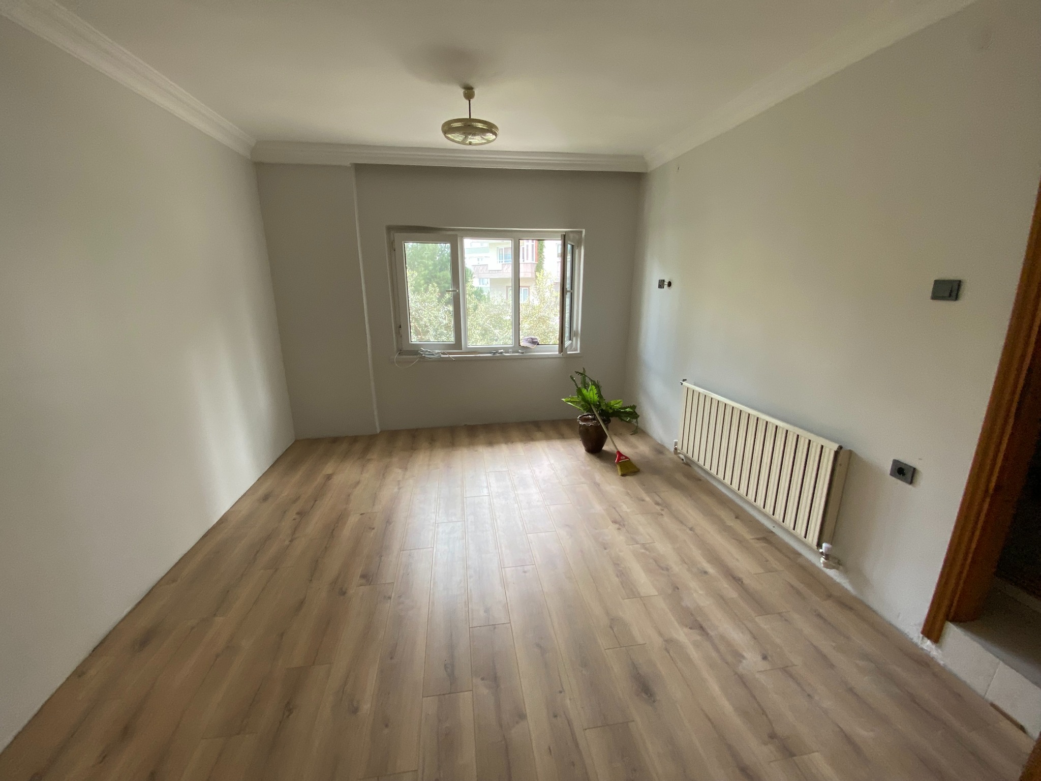 Jasne panele podłogowe ułożone w pokoju z grzejnikiem i rośliną w doniczce, widok na okno. Świeżo wykończone wnętrze po remoncie, neutralne kolory ścian.