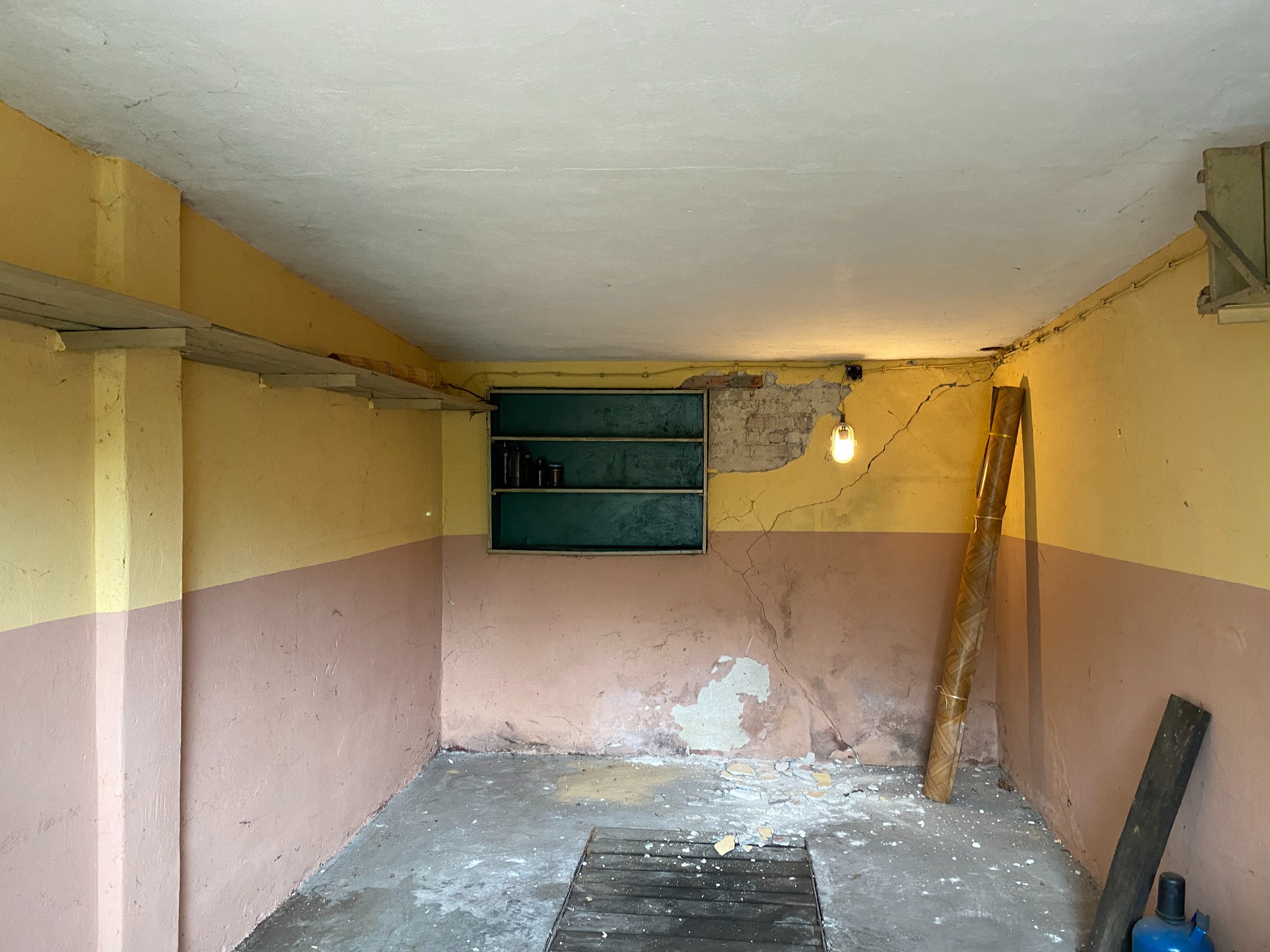 Wnętrze piwnicy z żółto-różowymi ścianami, widocznymi pęknięciami i odpadającym tynkiem. Na podłodze resztki gruzu i brudu. W rogu zwinięta wykładzina.