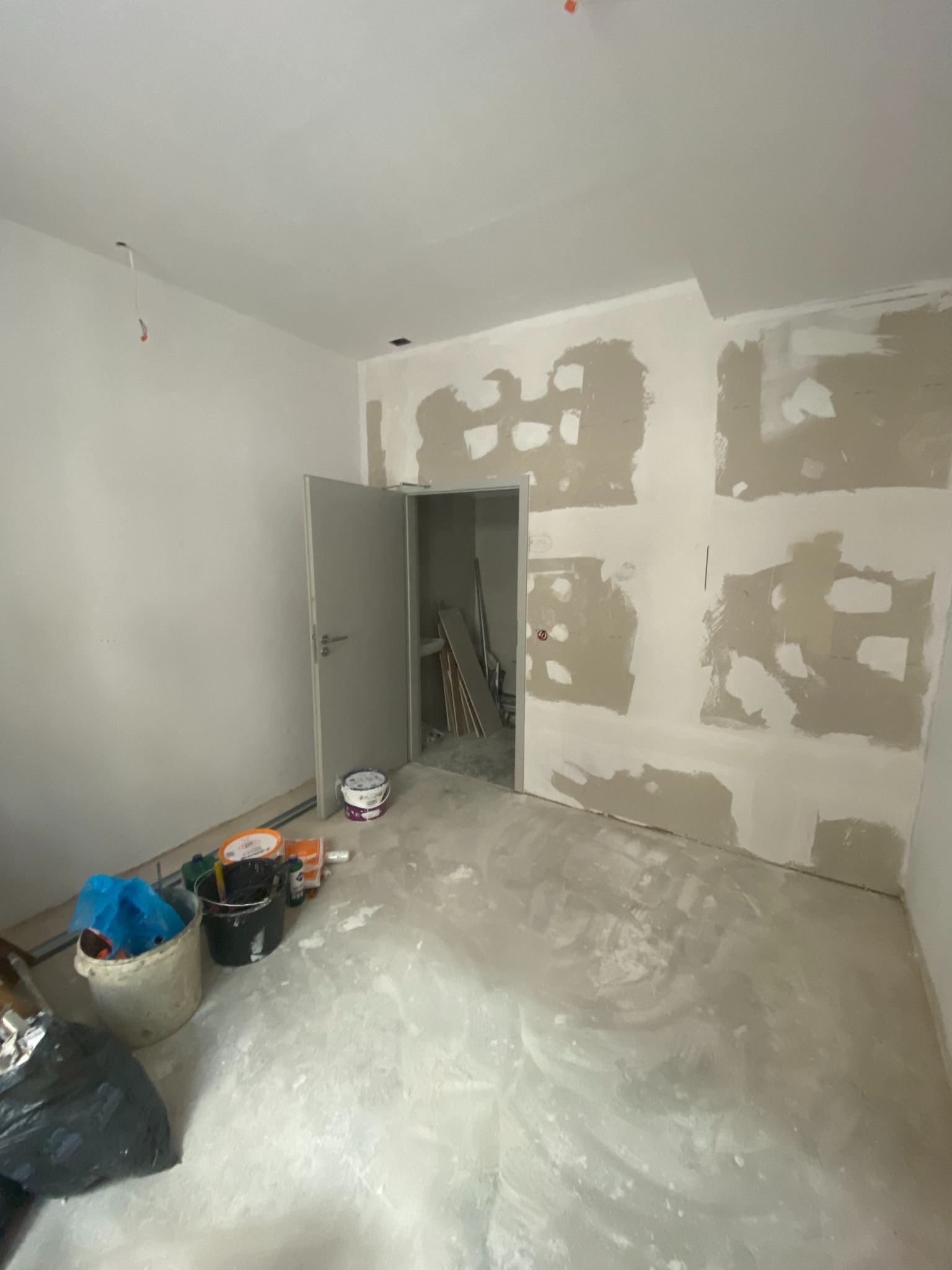 Wnętrze pokoju w trakcie remontu: ściany częściowo wyszpachlowane, widoczne narzędzia i materiały budowlane, otwarte drzwi do kolejnego pomieszczenia.