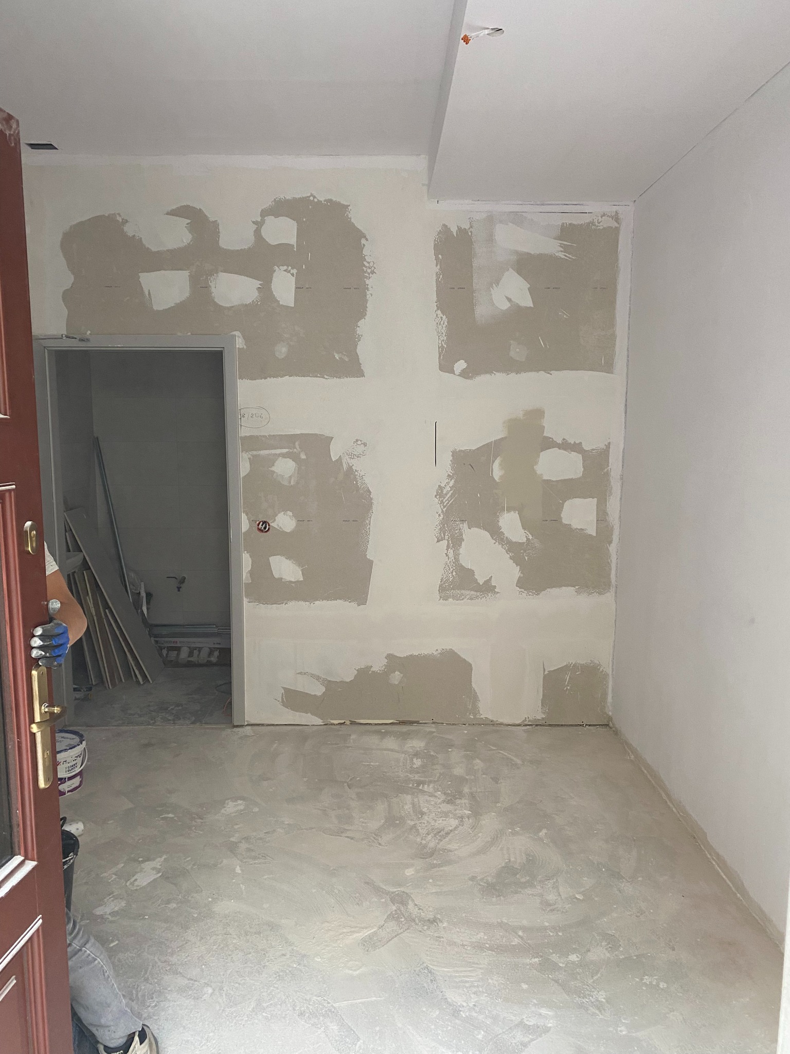 Pokój w trakcie szpachlowania, widoczne ślady po łatach na ścianach i podkład na podłodze. Otwarta, ciemna stolarka drzwiowa z widocznym wiadrem i narzędziami.