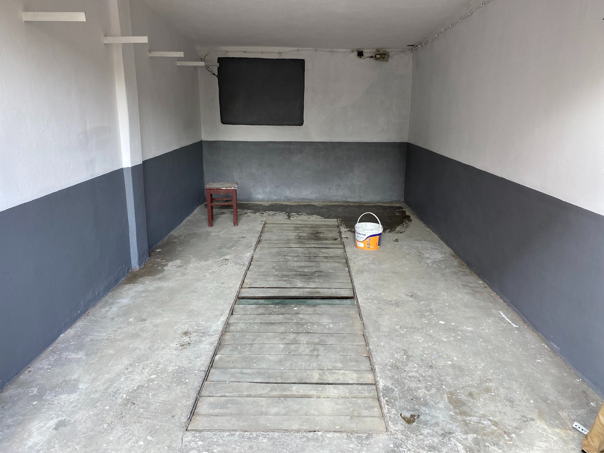 Wnętrze garażu po malowaniu: ściany dwukolorowe (szaro-białe), drewniana podłoga z otworem, wiadro z farbą, krzesło. Świeże malowanie, surowy, minimalistyczny styl.