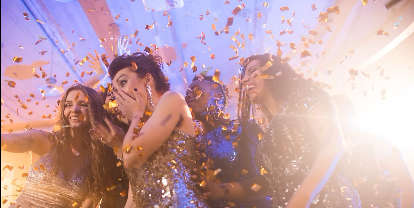 Rozbawione kobiety w błyszczących sukienkach świętują pod deszczem złotego konfetti, w tle jasne światła i fragmenty dekoracji sufitowej.