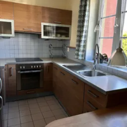 Jasna kuchnia z drewnianymi frontami, białymi płytkami na ścianie i oknem z wzorzystą zasłoną. Widoczny zlew, płyta indukcyjna i piekarnik.