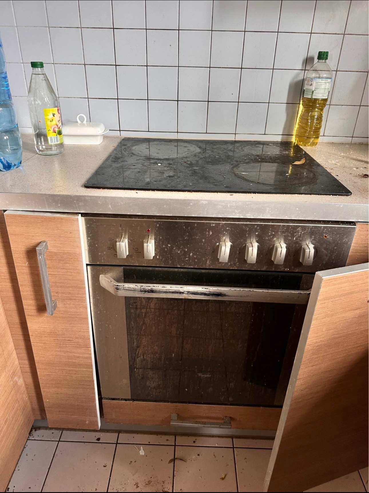Zabrudzona kuchnia z płytą grzewczą, piekarnikiem i butelkami. Widoczne plamy i resztki jedzenia. Zaniedbane wnętrze przed sprzątaniem. Otwarta szafka w dolnej części.