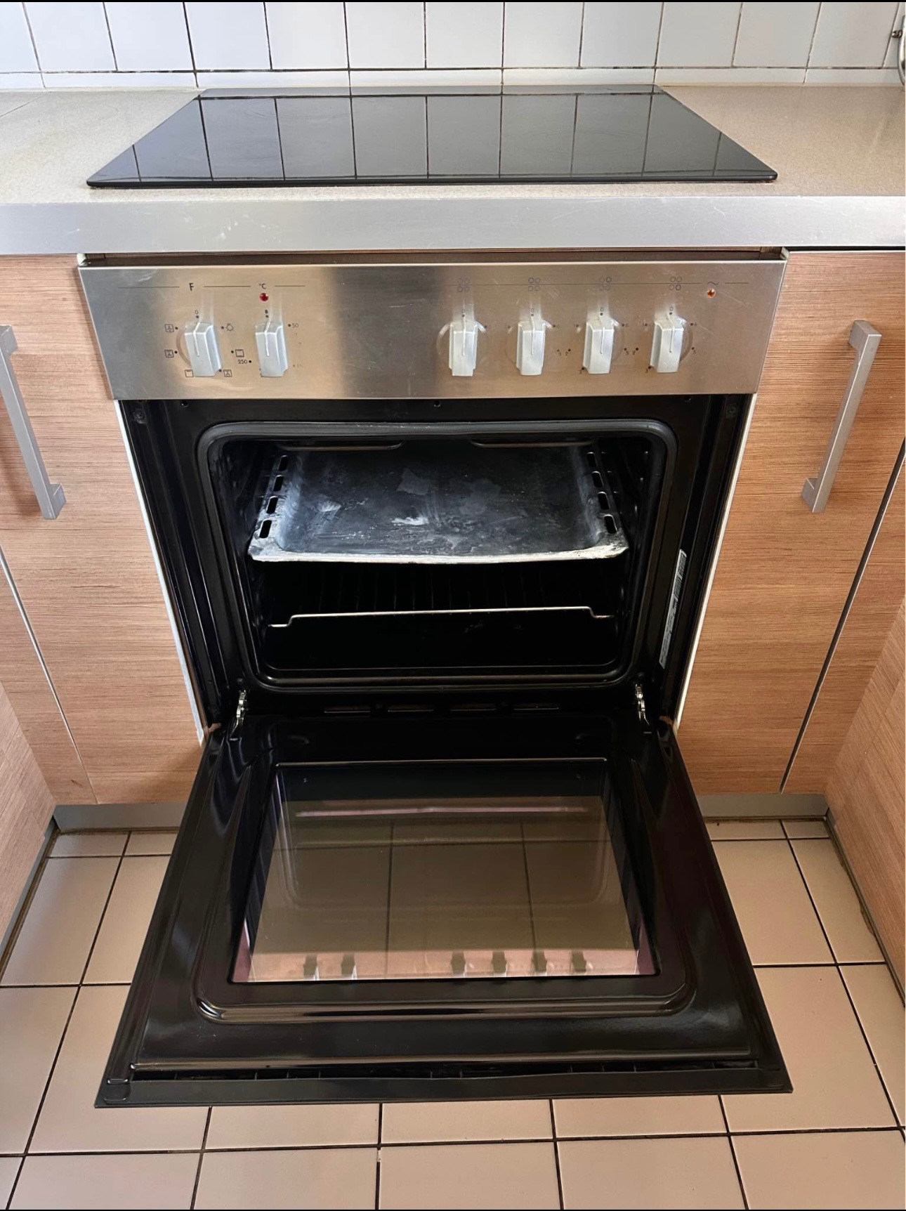 Otwarty, brudny piekarnik w zabudowie kuchennej z płytą grzewczą. Widoczne tłuste plamy i zabrudzenia wewnątrz piekarnika. Płytki na podłodze i ścianie.