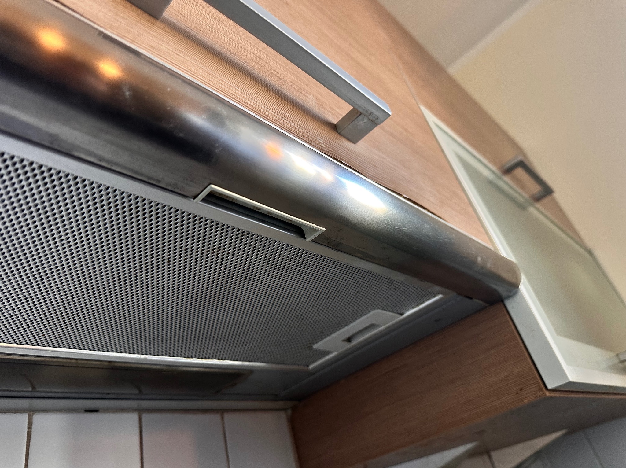 Kuchenny okap podszafkowy ze srebrną listwą i metalową siatką filtracyjną, widziany z dołu pod kątem. Widoczne płytki i drewniane szafki.