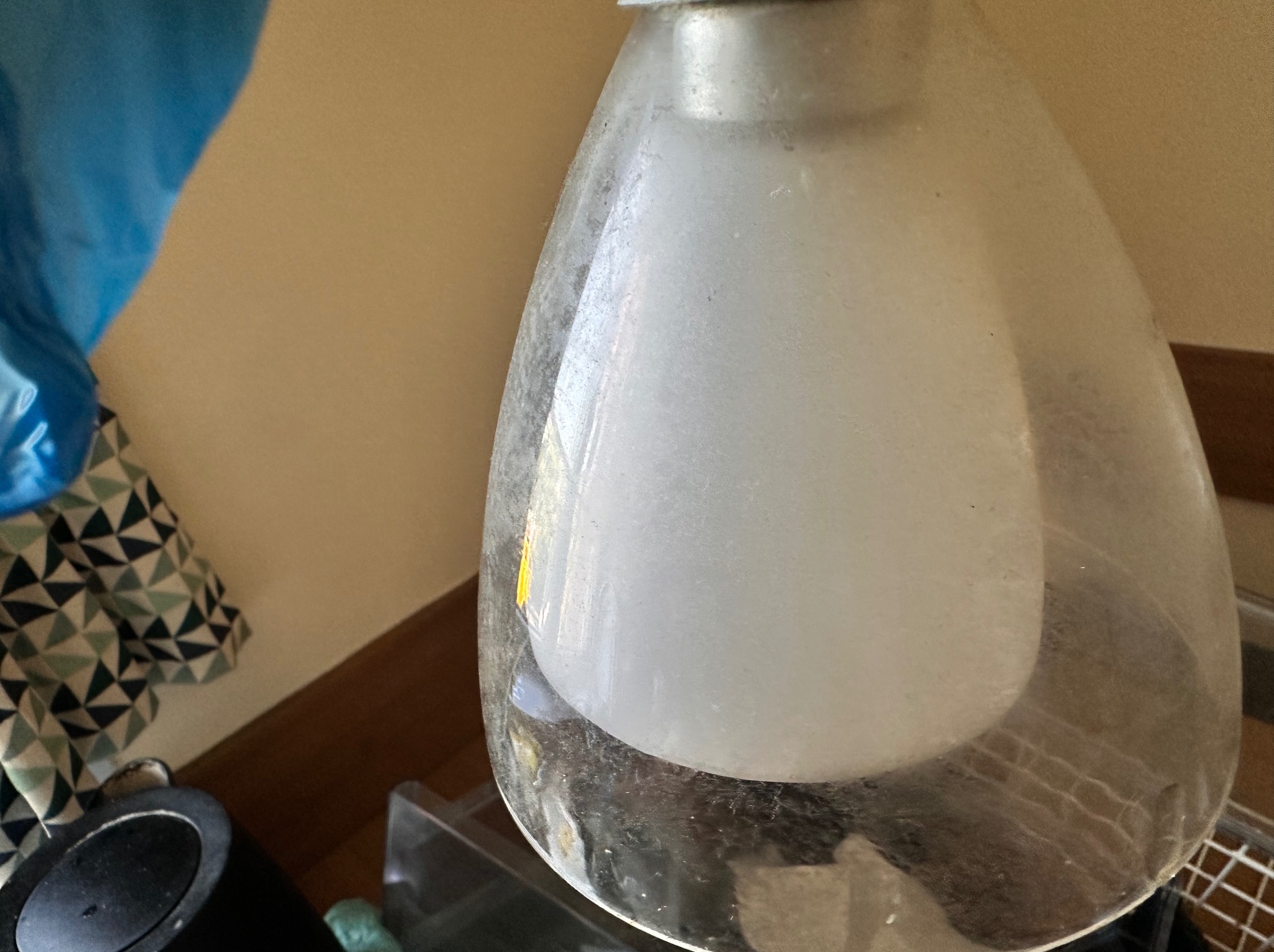 Brudna lampa wisząca z osadem i kurzem, widoczne zabrudzenia na kloszu, w tle ściana i fragmenty wyposażenia wnętrza.