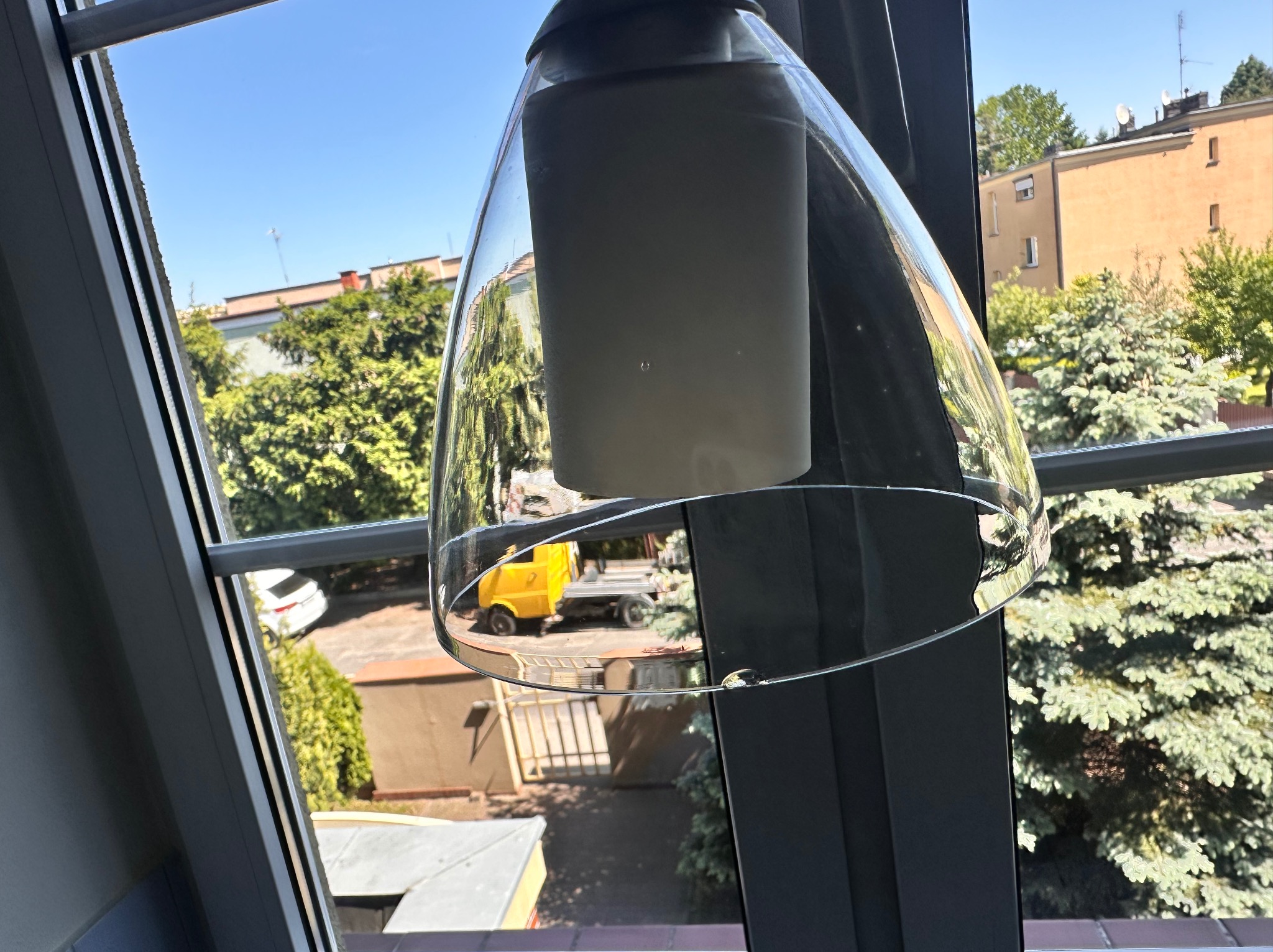 Lampa wisząca z przezroczystym kloszem, widok z okna na budynki i zieleń. Na zewnątrz żółty wózek transportowy. Słoneczny dzień.