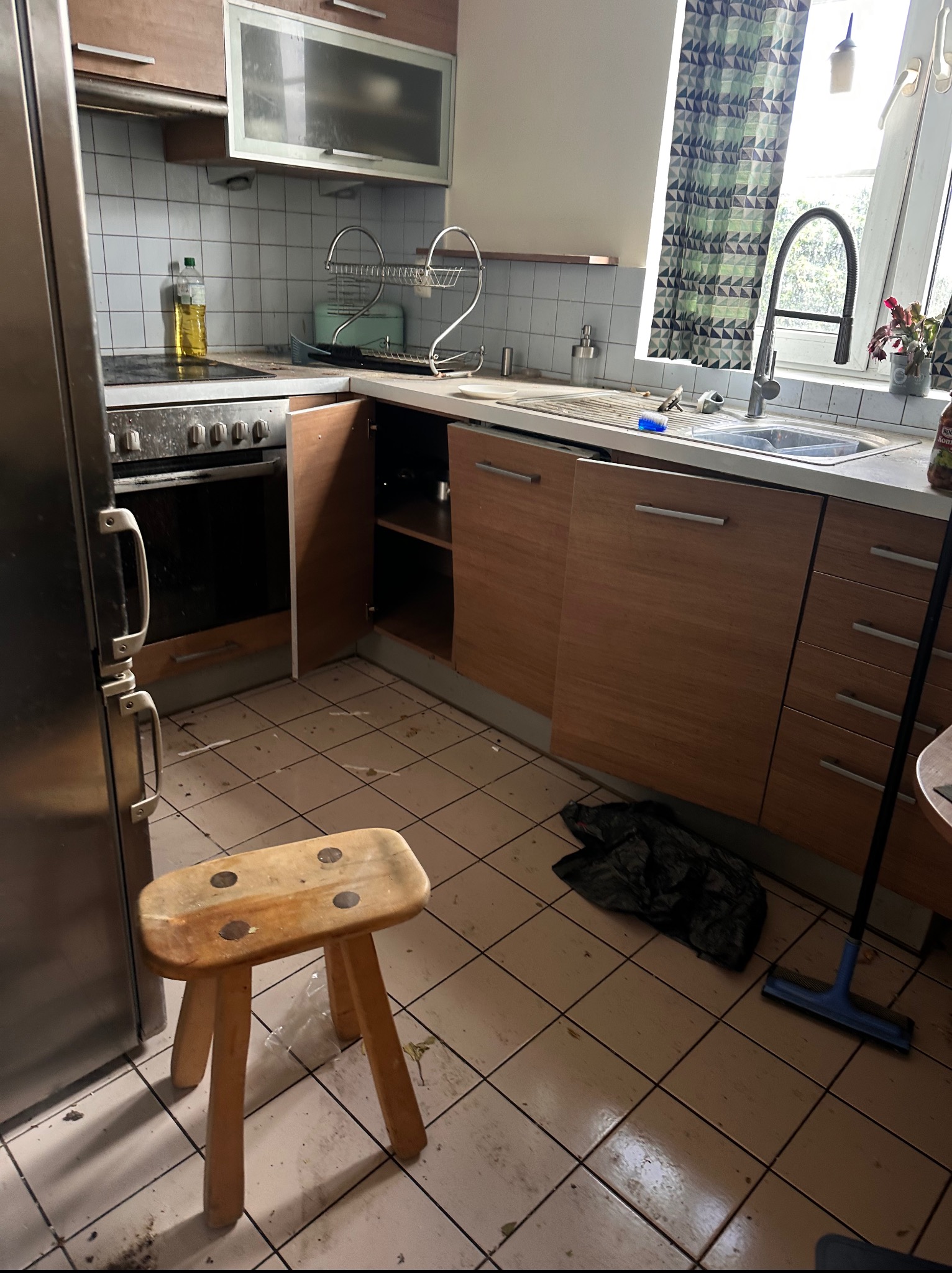 Zaniedbana kuchnia z brudnym piekarnikiem, zlewem i podłogą w płytkach. Drewniany stołek i mop leżą na podłodze. Widoczne otwarte szafki i okno z zasłoną.