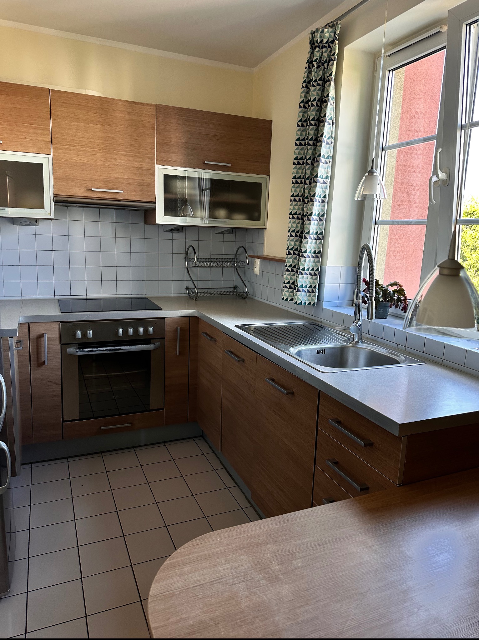 Jasna kuchnia z drewnianymi frontami, białymi płytkami na ścianie i oknem z wzorzystą zasłoną. Widoczny zlew, płyta indukcyjna i piekarnik.