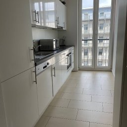 SUCCESS KID SPÓŁKA Z OGRANICZONĄ ODPOWIEDZIALNOŚCIĄ - Biała, nowoczesna kuchnia w mieszkaniu z widokiem na budynek przez okno. Umyte płytki podłogowe i błyszczące fronty szafek.