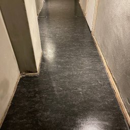 SUCCESS KID SPÓŁKA Z OGRANICZONĄ ODPOWIEDZIALNOŚCIĄ - Błyszcząca, ciemna podłoga po czyszczeniu w korytarzu z szarymi ścianami i białymi drzwiami. Widoczne listwy przypodłogowe i narożny filar.