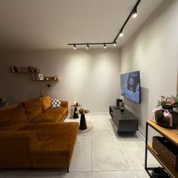 SUCCESS KID SPÓŁKA Z OGRANICZONĄ ODPOWIEDZIALNOŚCIĄ - Salon z narożnikiem w odcieniach brązu, oświetleniem szynowym i elementami dekoracyjnymi. Neutralne kolory ścian i podłogi, minimalistyczny styl.