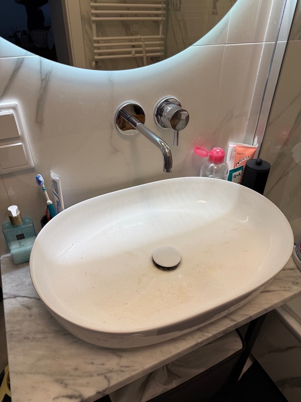 Owalna umywalka nablatowa z chromowaną armaturą na marmurowym blacie, widoczne drobne zabrudzenia i włosy. W tle podświetlane lustro i grzejnik drabinkowy.