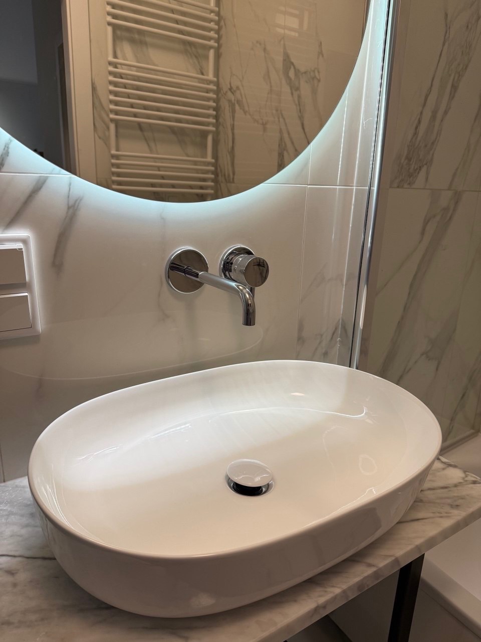 Biała, owalna umywalka nablatowa z chromowaną baterią na marmurowym blacie. W tle lustro z podświetleniem i grzejnik drabinkowy.