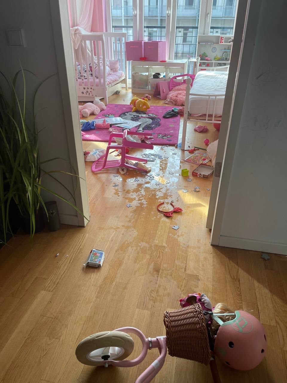 Rozrzucone zabawki, puzzle i ubrania na podłodze w pokoju dziecięcym z różowymi akcentami, widok z perspektywy wejścia.