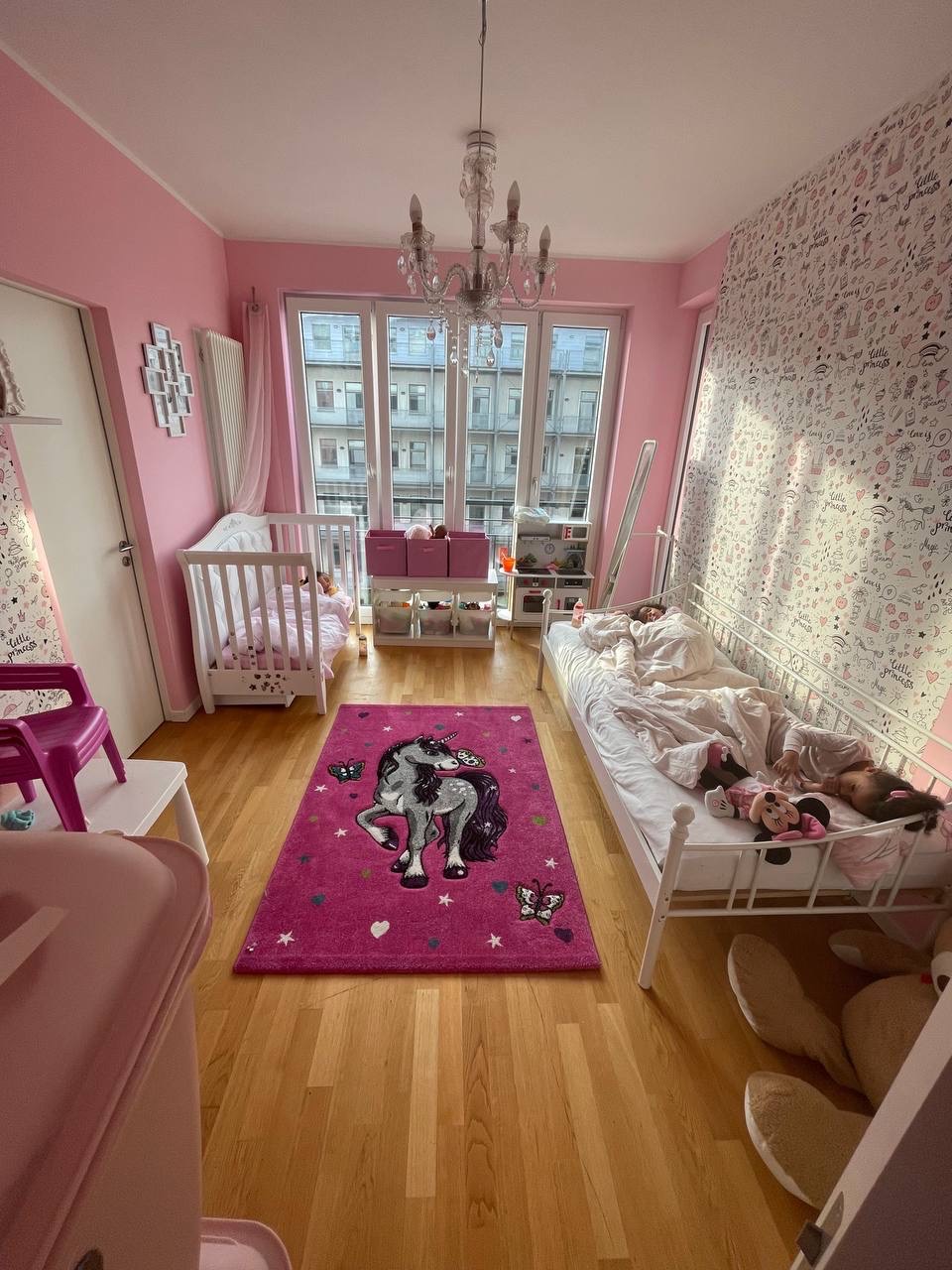 Różowy pokój dziecięcy z dwoma łóżkami, dywanem z jednorożcem i oknem z widokiem na miasto. Na jednym łóżku śpi dziewczynka.