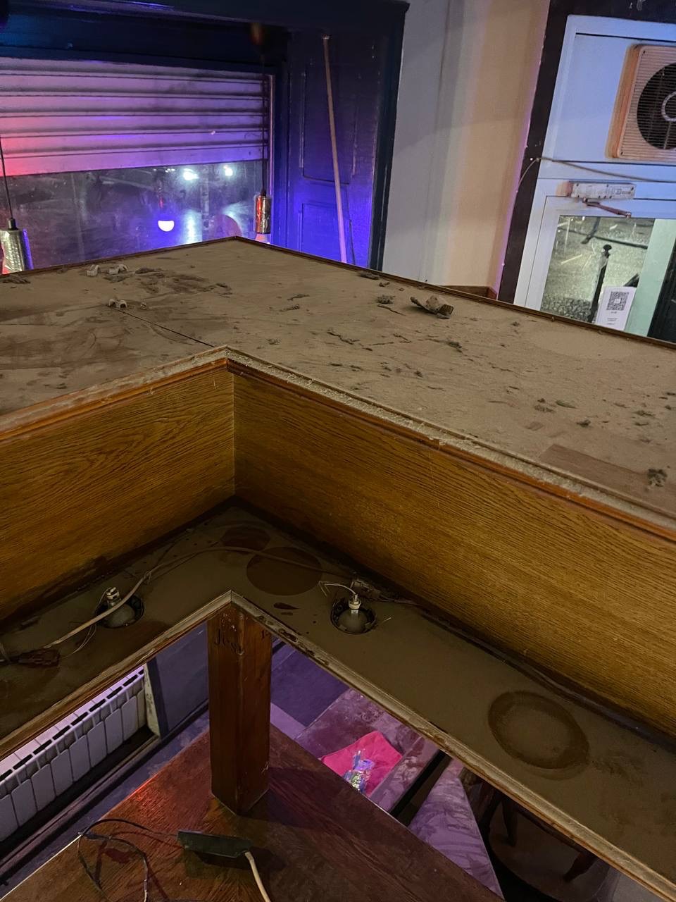 Zabrudzony, drewniany bar w lokalu z widocznymi śladami kurzu i zaniedbania. W tle okno z roletą i klimatyzator.