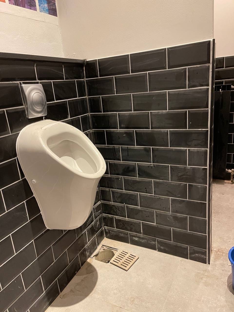 Biały pisuar na ścianie z czarnych płytek z białą fugą, widoczny odpływ pod pisuarzem i fragment podłogi. Ujęcie z dołu, ciemne oświetlenie.