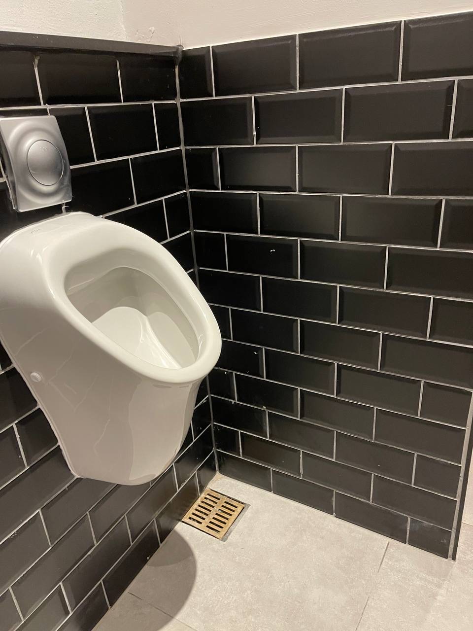 Biały pisuar ceramiczny na czarnych, błyszczących płytkach w nowoczesnej toalecie. Widoczna kratka odpływowa w podłodze i przycisk spłukiwania.