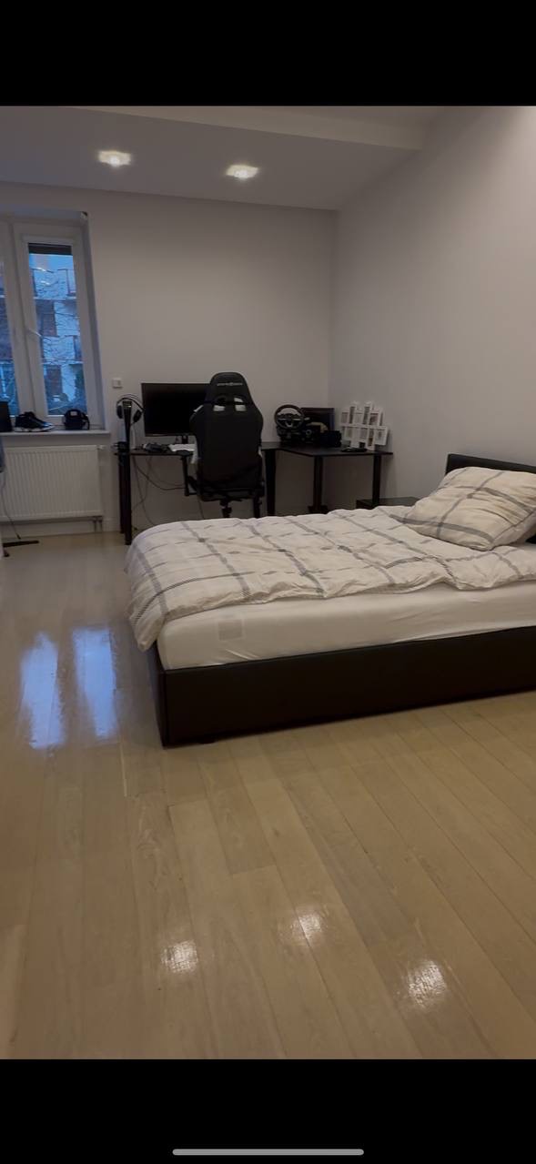 Pokój z łóżkiem, biurkiem gamingowym i oknem. Jasne panele podłogowe, stonowana kolorystyka ścian. Minimalistyczny wystrój, widoczne akcesoria gamingowe.