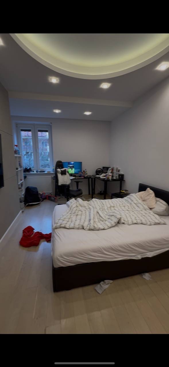 Niedbale posłane łóżko w pokoju z biurkiem i komputerem. Na podłodze leżą ubrania. Widok na okno zza którego widać budynki.