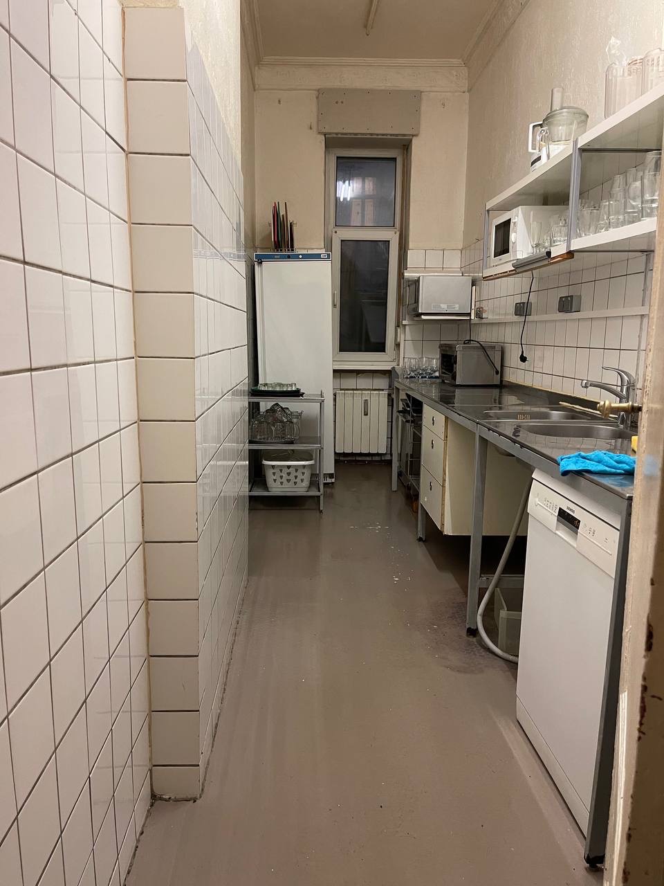 Wnętrze kuchni z białymi kafelkami na ścianach i podłodze. Widoczny zlewozmywak, półki z naczyniami, lodówka i okno. Minimalistyczny styl, brak dekoracji.