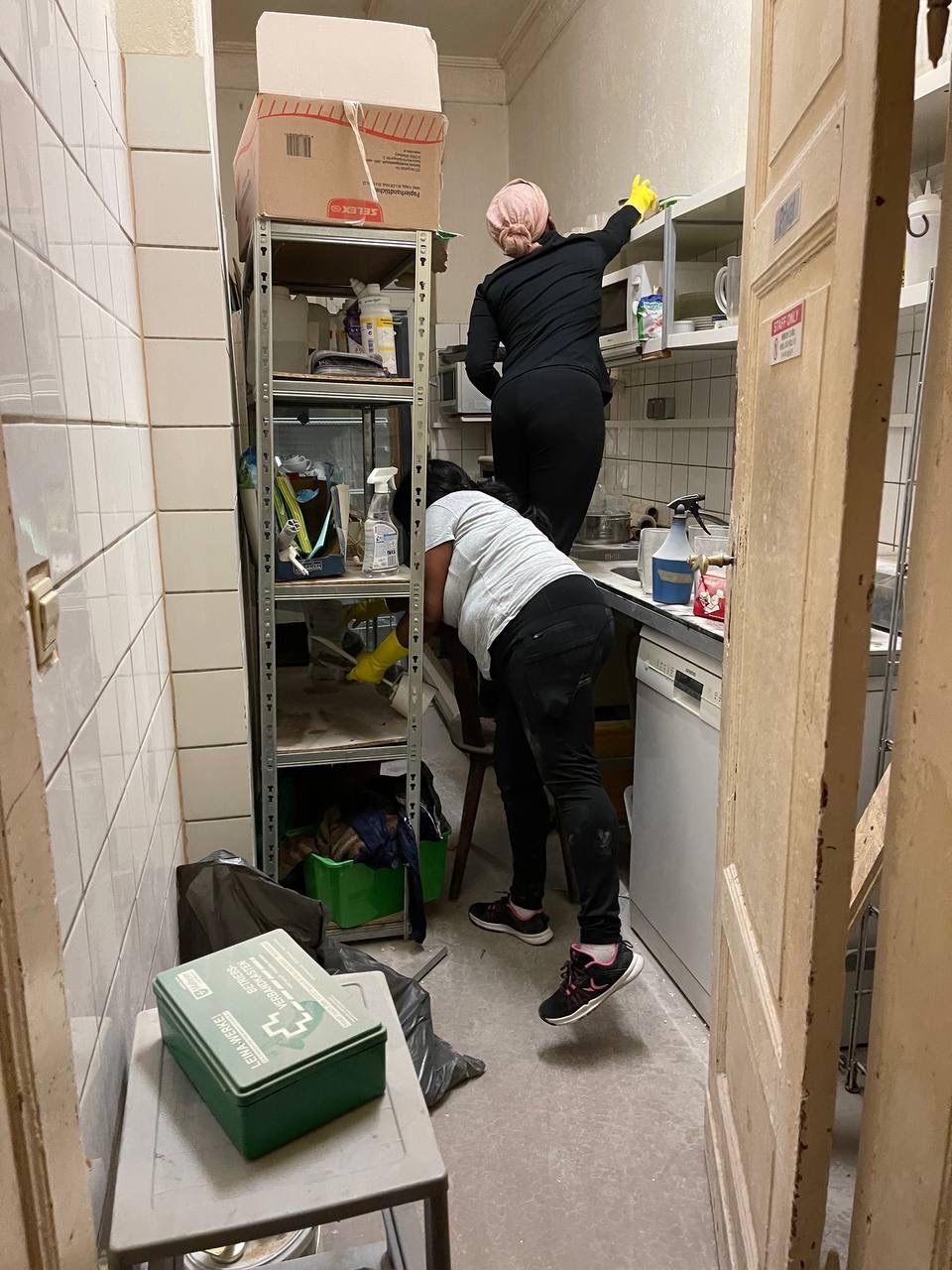 Dwie osoby sprzątają kuchnię z białymi kafelkami. Jedna czyści szafki, druga stoi na stołku przy półce. Widoczna apteczka i środki czystości.