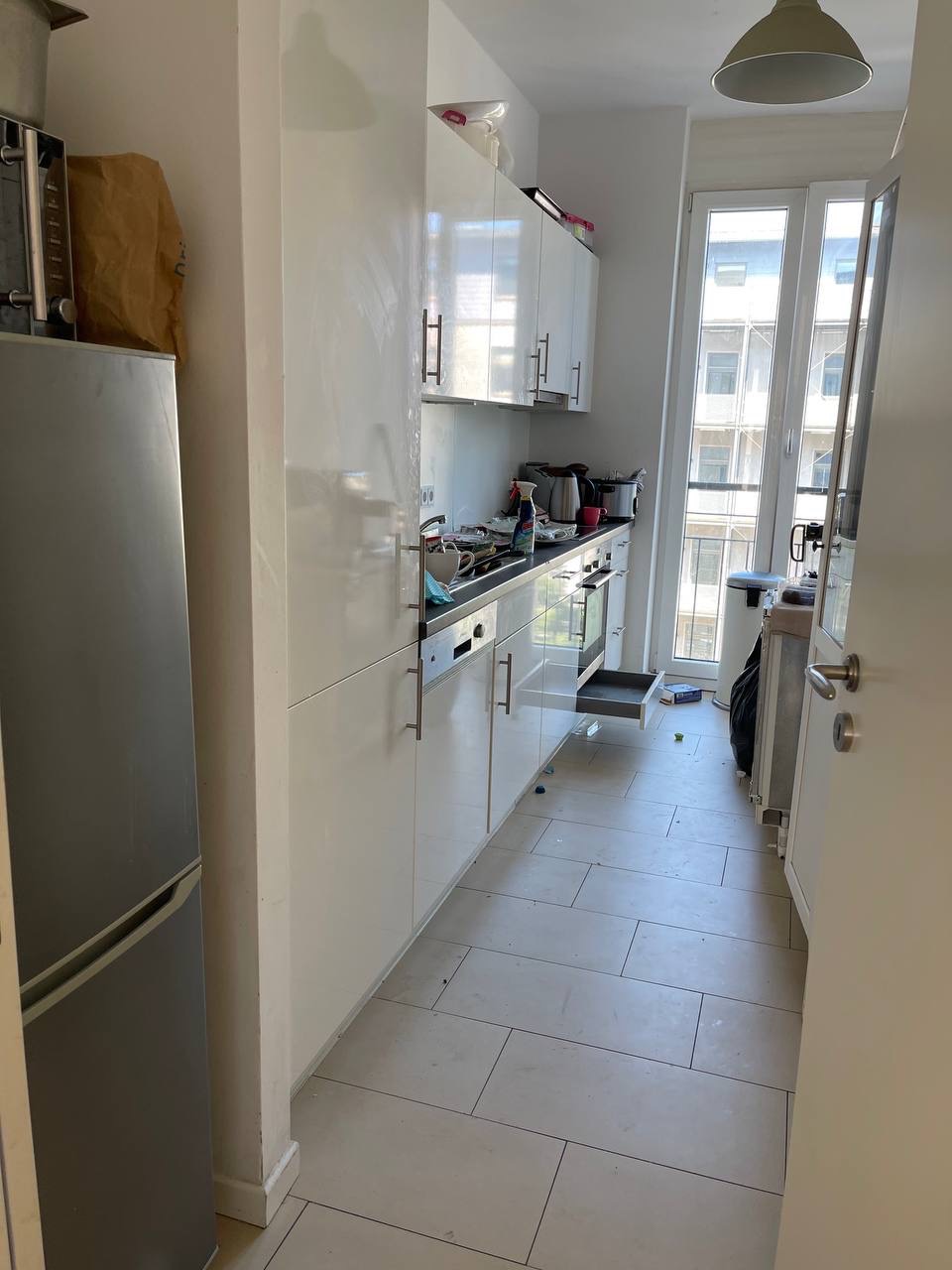 Widok na zagraconą kuchnię z białymi szafkami, srebrną lodówką i otwartymi drzwiami balkonowymi. Na blacie widoczne naczynia i butelka ze środkiem czyszczącym.