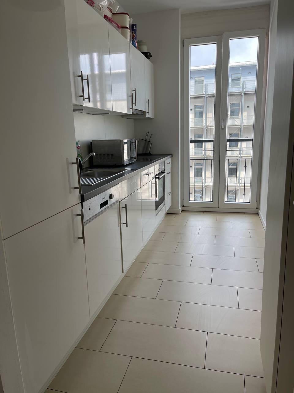 Biała, nowoczesna kuchnia w mieszkaniu z widokiem na budynek przez okno. Umyte płytki podłogowe i błyszczące fronty szafek.