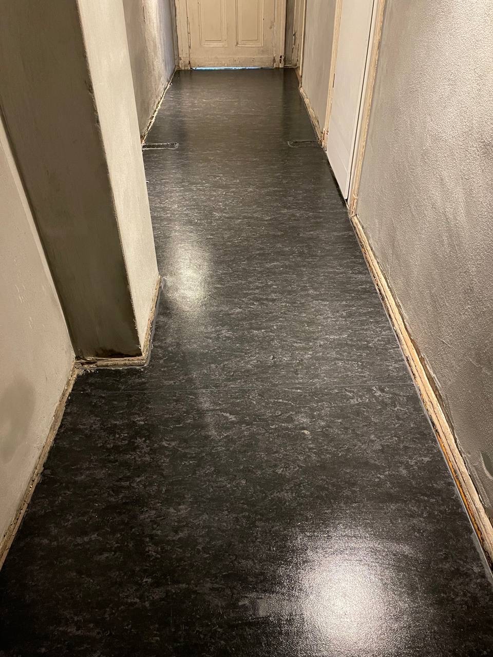 Błyszcząca, ciemna podłoga po czyszczeniu w korytarzu z szarymi ścianami i białymi drzwiami. Widoczne listwy przypodłogowe i narożny filar.