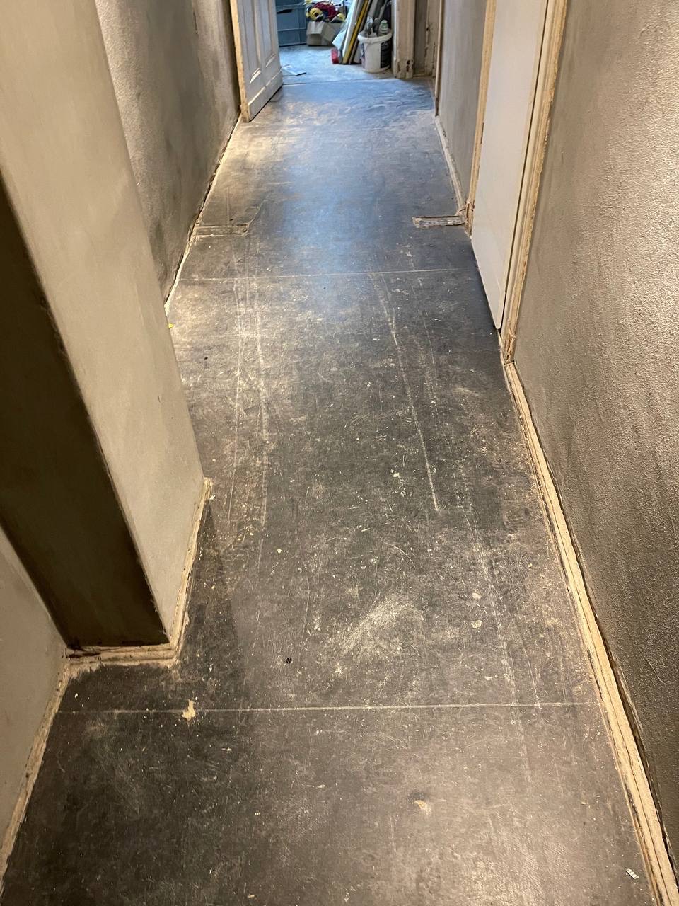 Korytarz po remoncie z szarą podłogą, ściany w surowym tynku i widoczne ślady po pracach budowlanych. W tle otwarte drzwi i pomieszczenie z narzędziami.