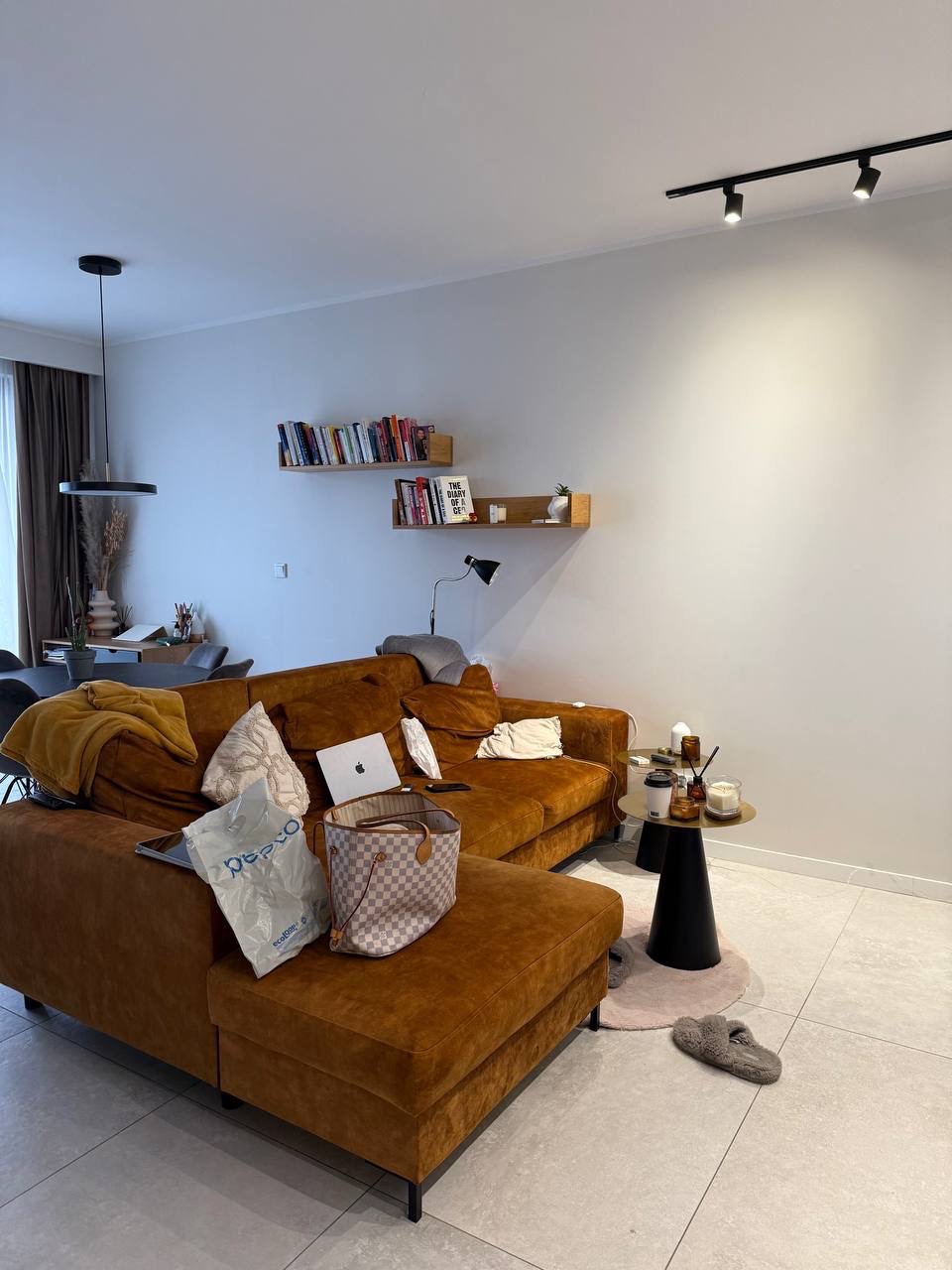 Salon z brązową sofą narożną, stolikiem kawowym i półkami na ścianie. Widoczne osobiste przedmioty: laptop, torba, książki, dodające wnętrzu autentycznego charakteru.