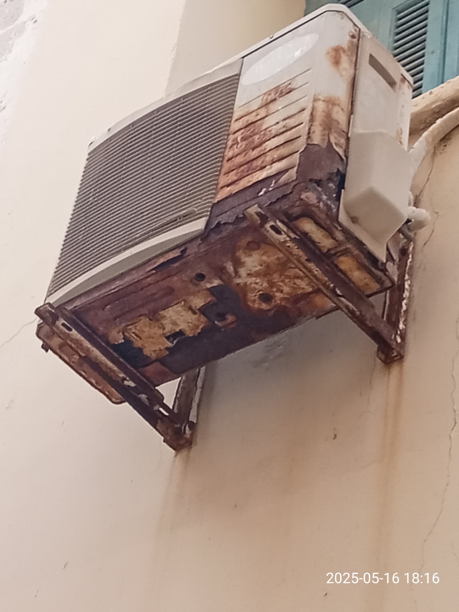 Zardzewiała, stara klimatyzacja zewnętrzna na ścianie budynku. Widoczne ślady korozji i uszkodzenia obudowy. Niewyraźne okno w tle.