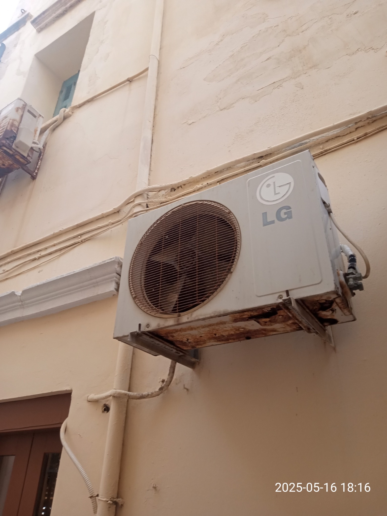 Zewnętrzna jednostka klimatyzacji LG zamontowana na ścianie budynku. Widoczne ślady korozji na obudowie i wspornikach. Dodatkowa jednostka klimatyzacji w tle.