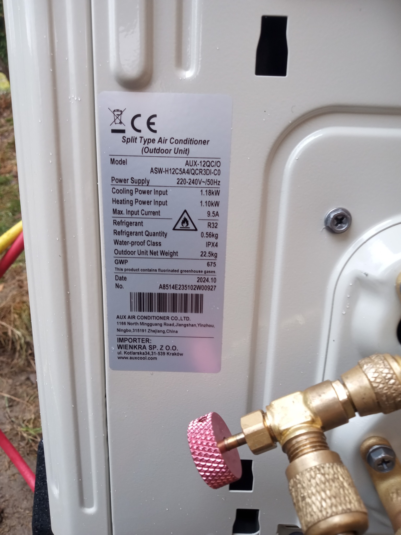 Zewnętrzna jednostka klimatyzacji AUX z tabliczką znamionową, na której widoczne są dane techniczne, podłączona do manometru podczas serwisu. Widoczne ślady wilgoci.
