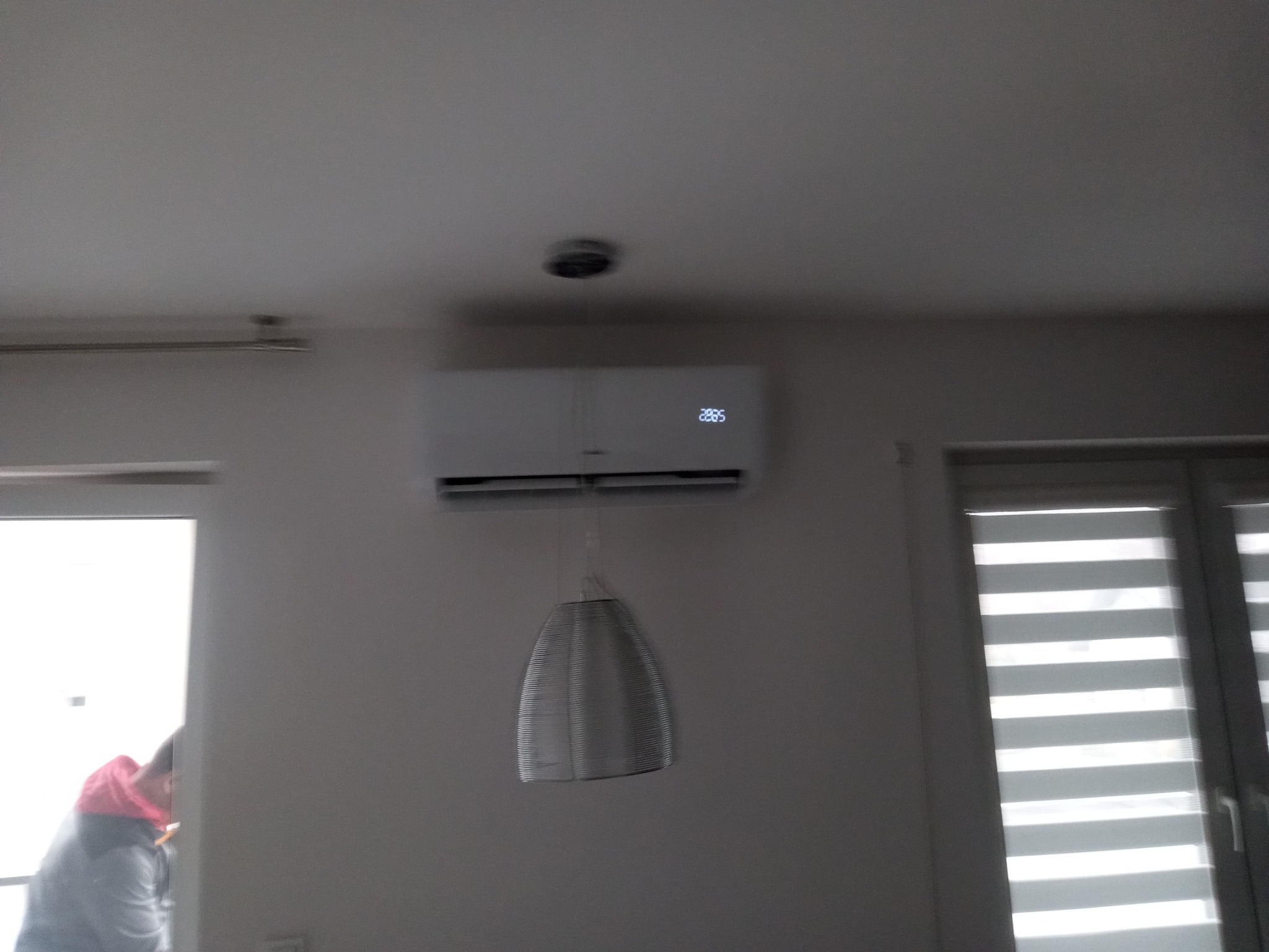 Biały klimatyzator ścienny z wyświetlaczem temperatury, zawieszony nad lampą w nowoczesnym wnętrzu. Widoczny fragment osoby w lewym dolnym rogu.