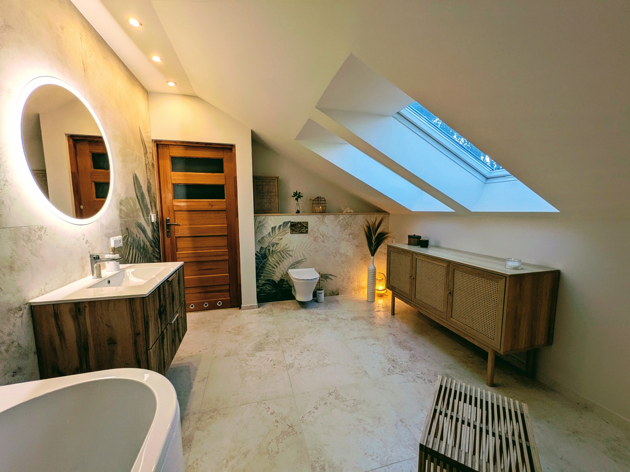 Stylowa łazienka na poddaszu w Łodzi z drewnianymi akcentami: szafka pod umywalką, komoda z rattanowymi frontami i drewniane drzwi. Uwagę przykuwa okrągłe lustro z podświetleniem LED.