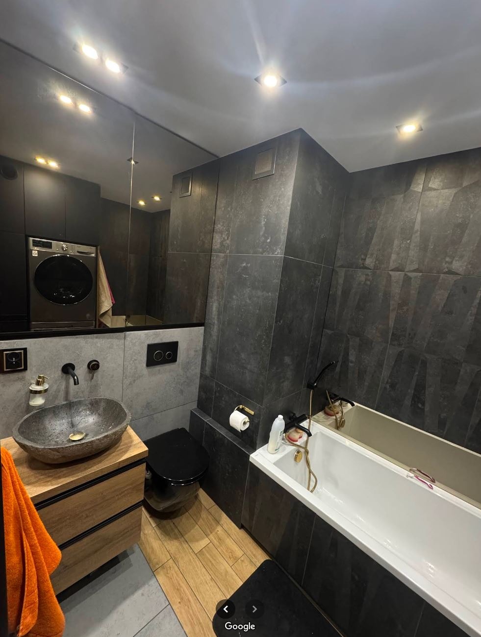 Nowoczesna łazienka z kamienną umywalką, czarną armaturą i płytkami imitującymi kamień. Widoczna pralka w zabudowie, drewniana podłoga i minimalistyczne oświetlenie.