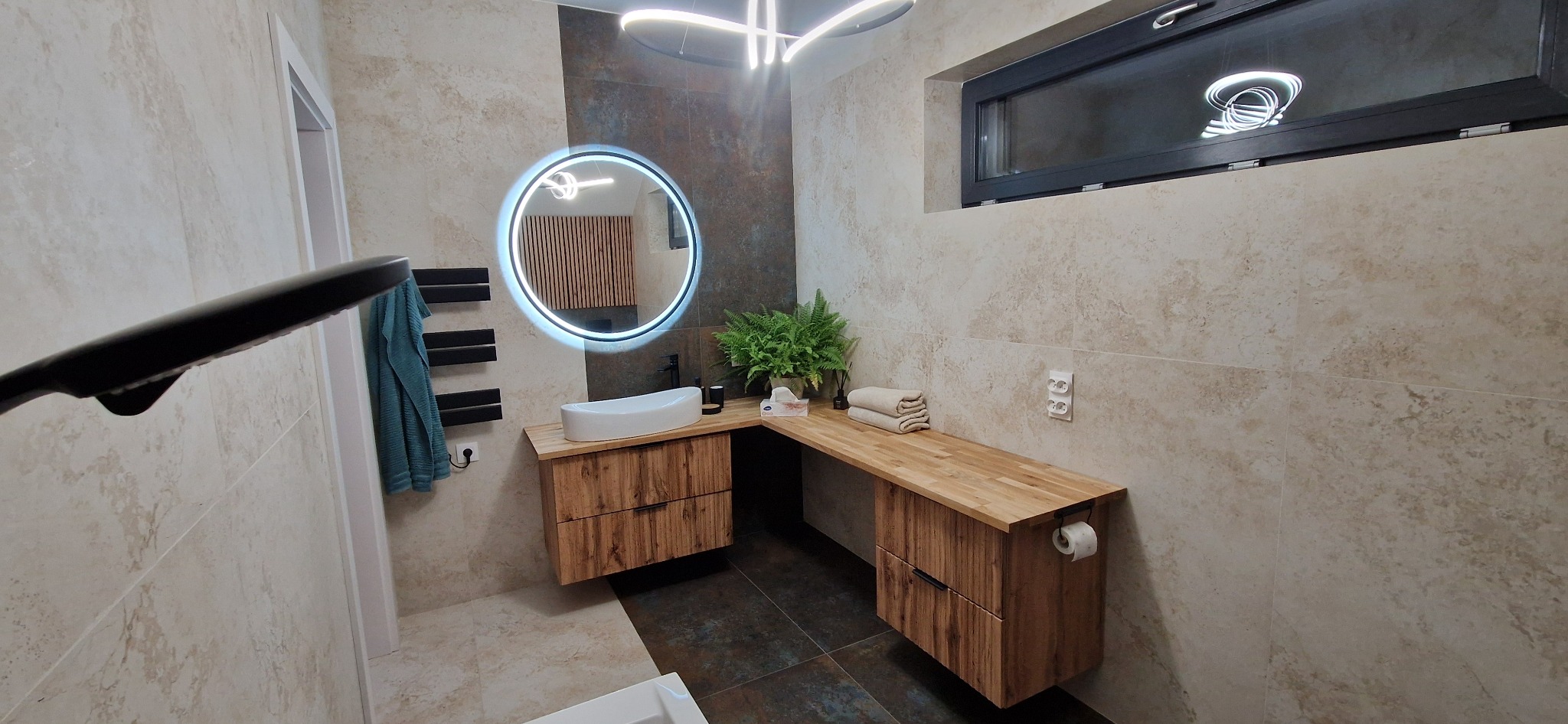 Nowoczesna łazienka z drewnianą zabudową umywalki, okrągłym lustrem z podświetleniem LED i minimalistycznym oświetleniem sufitowym.