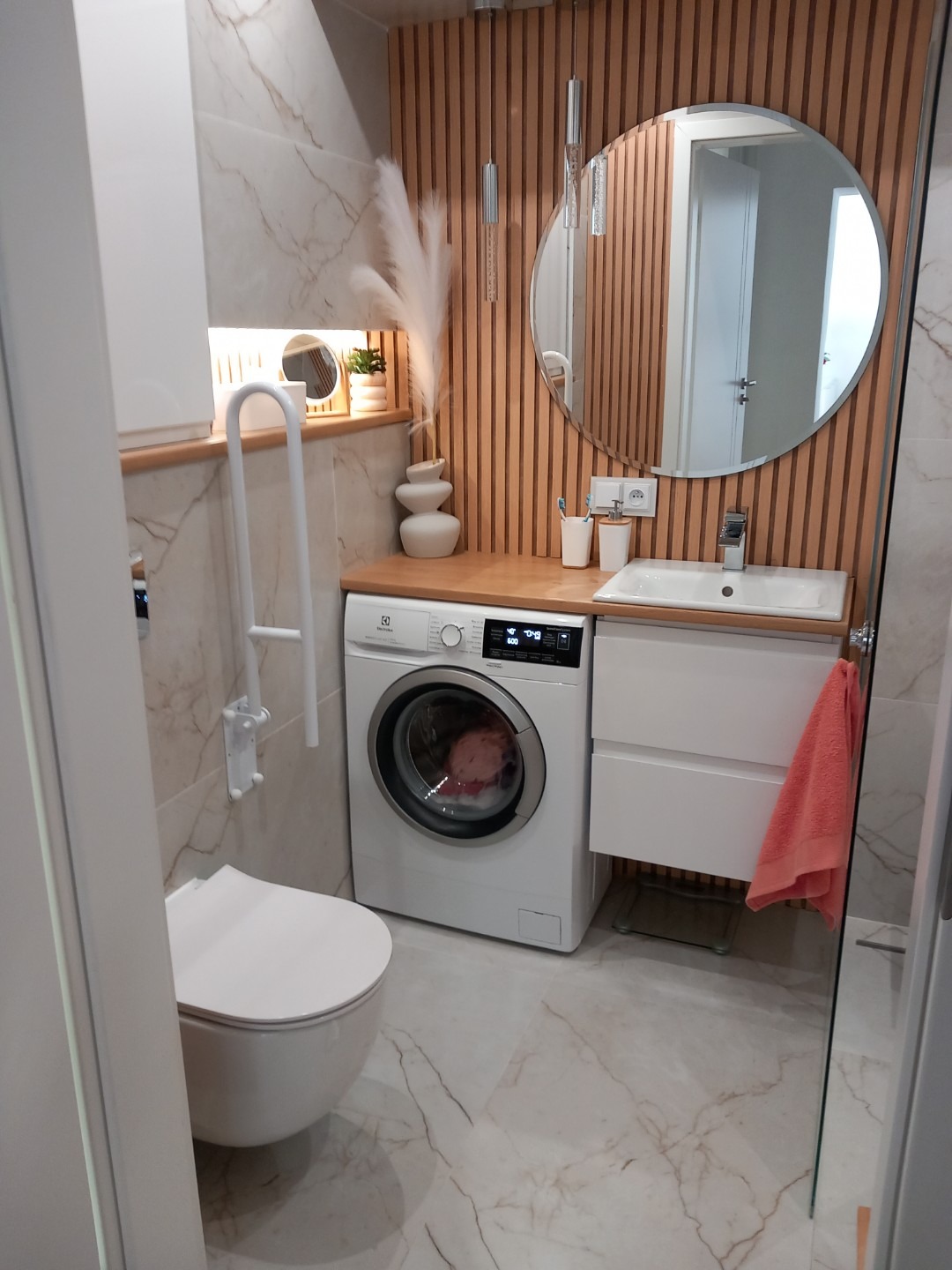 Funkcjonalna łazienka z pralką pod blatem, okrągłym lustrem i drewnianymi lamelami na ścianie. Umywalka w zabudowie, toaleta podwieszana. Marmurowe płytki na podłodze.