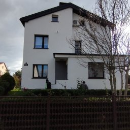 Woj-Bud - Dwupiętrowy dom z białą elewacją i ciemnym dachem, widoczny zza ogrodzenia. Na ścianie numer '73a'. Drzewo bez liści po prawej. Pochmurne niebo w tle.