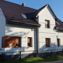 Dwukondygnacyjny dom z ciemnobrązowym dachem i jasną elewacją, z elementami dekoracyjnymi imitującymi drewno. Widoczne okna z roletami i czarne ogrodzenie.