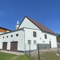 Woj-Bud - Biały budynek z brązowym dachem, malowany przez pracownika na rusztowaniu. W tle widoczna wieża kościoła i błękitne niebo.