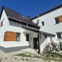 Biały dom z brązowym dachem i drewnianymi akcentami elewacyjnymi, widziany z zewnątrz w słoneczny dzień. Widoczne krzewy róż przed budynkiem.