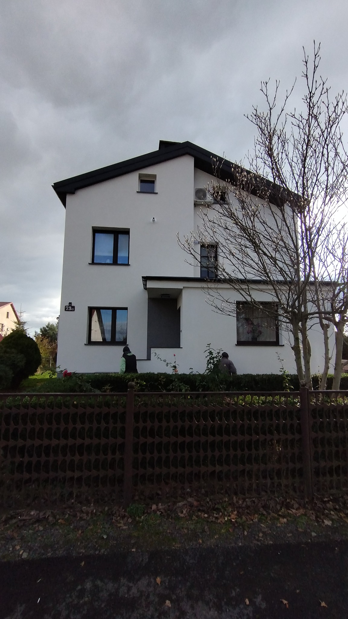 Dwupiętrowy dom z białą elewacją i ciemnym dachem, widoczny zza ogrodzenia. Na ścianie numer '73a'. Drzewo bez liści po prawej. Pochmurne niebo w tle.