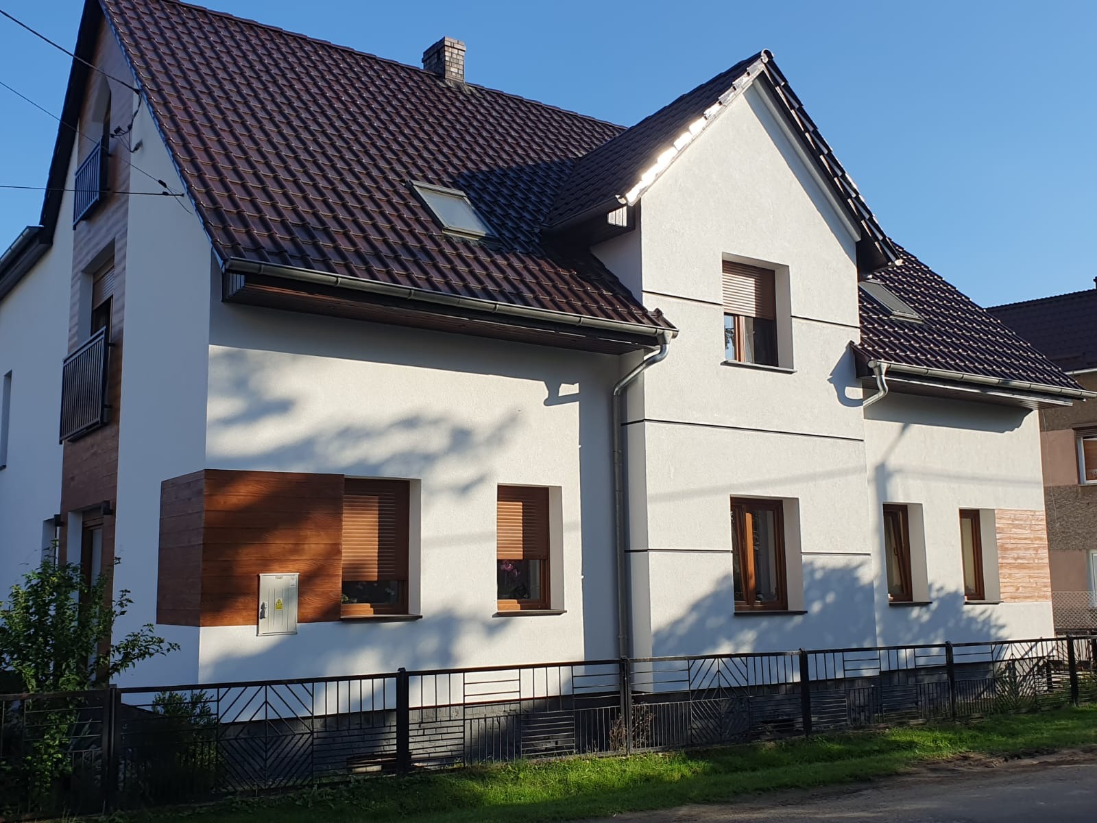 Dwukondygnacyjny dom z ciemnobrązowym dachem i jasną elewacją, z elementami dekoracyjnymi imitującymi drewno. Widoczne okna z roletami i czarne ogrodzenie.