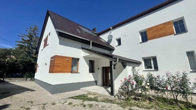 Biały dom z brązowym dachem i drewnianymi akcentami elewacyjnymi, widziany z zewnątrz w słoneczny dzień. Widoczne krzewy róż przed budynkiem.