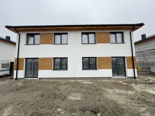 Dwukondygnacyjny, biały dom bliźniak z drewnianymi panelami elewacyjnymi i czarnymi ramami okiennymi, zabezpieczonymi folią. Na pierwszym planie błotniste podłoże.